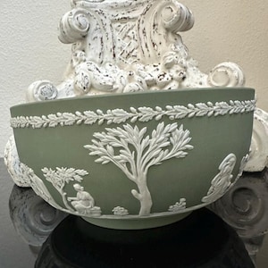 May include: A sage green ceramic bowl with white relief detailing. The bowl features a decorative border and scenes of trees and figures. The bowl is set on a black surface, with a white, ornate base behind it.
