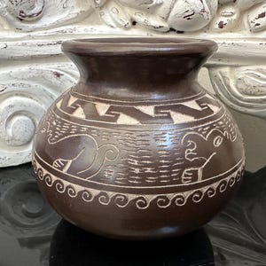 May include: A small, round, brown ceramic vase with a dark rim and intricate carved designs. The vase features a repeating geometric pattern and stylized animal figures in a lighter color, creating a contrast against the dark brown background.