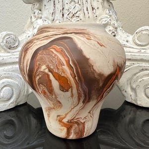 May include: A handcrafted ceramic vase with a unique marbled design. The vase features a blend of brown, orange, and cream colors, creating a swirling pattern. The vase has a rounded body and a slightly flared rim, suitable for home decor.