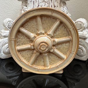 May include: A ceramic wagon wheel-shaped decorative piece in shades of beige and brown. The wheel features eight spokes radiating from a central hub. The piece is displayed on a reflective black surface, with a white, ornate background.