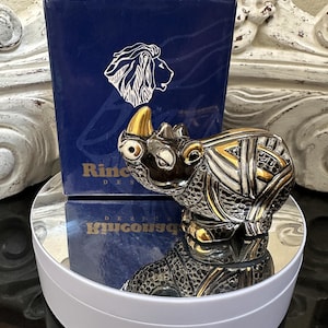 May include: A silver and gold rhino figurine with a geometric design, displayed on a white rotating stand. A navy blue box with a lion emblem and the word "Rinconada" is behind the rhino.