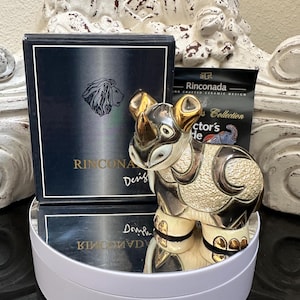 May include: A ceramic rhinoceros figurine with gold accents, displayed on a white platform. The figurine is next to a black box with a lion logo and the text "Rinconada Design". A collector's card is also visible.