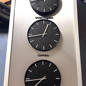 May include: A rectangular wall-mounted display featuring three circular black clocks with silver hands and markers. The clocks are labeled United Kingdom, Germany, and Japan in black text on a brushed silver metallic background within a silver frame.