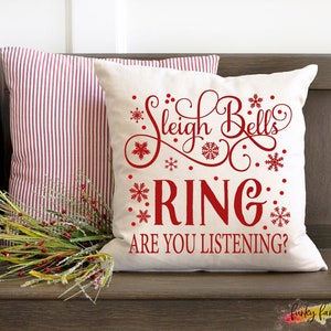 May include: Two decorative Christmas pillows on a wooden bench. One pillow is cream-colored with red text that reads "Sleigh Bells Ring, Are You Listening?" and snowflake accents. The other pillow has red and white stripes.