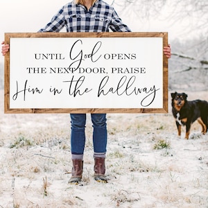 May include: A rectangular wooden sign with the text "Until God opens the next door, praise Him in the hallway" in black script. The sign is held by a person in a snowy field, with a dog in the background.