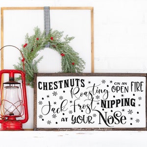 May include: A rustic Christmas-themed display featuring a red lantern, a framed sign with the text "Chestnuts Roasting on an Open Fire," and a green wreath with red berries. The sign is framed in dark wood.