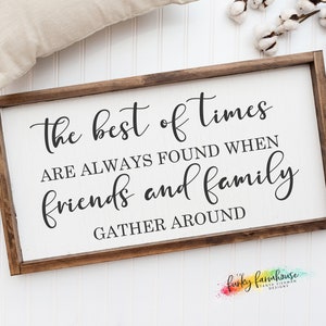 Op de afbeelding: Een rechthoekig houten bord met de tekst "The best of times are always found when friends and family gather around" in zwart schrift. Het bord heeft een witte achtergrond en een donkerbruine lijst. Een beige kussen en katoentakken op de achtergrond.