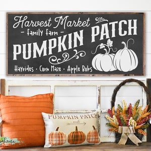Könnte beinhalten: Eine herbstliche Dekoration mit einem Tafelschild mit der Aufschrift "Harvest Market - Pumpkin Patch" und Kürbisillustrationen. Darunter ein orangefarbenes Kissen, ein Kissen mit Kürbismuster und ein Korb mit Trockenblumen.