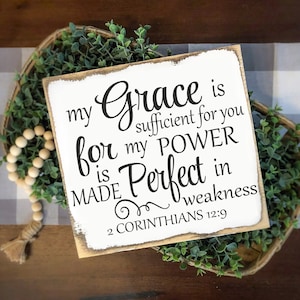 Op de afbeelding: Een vierkant, wit houten bord met de tekst "my Grace is sufficient for you, for my POWER is MADE Perfect in weakness 2 CORINTHIANS 12:9" in zwart schrift. Het bord staat in een gevlochten mand met groen.