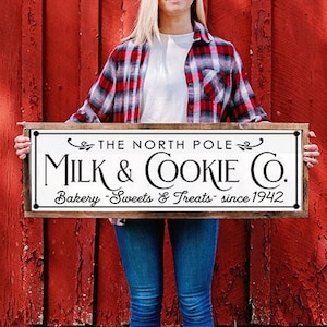 May include: A rectangular wooden sign with the text "THE NORTH POLE MILK & COOKIE CO. Bakery Sweets & Treats since 1942" in black and white. The sign is held in front of a red barn.