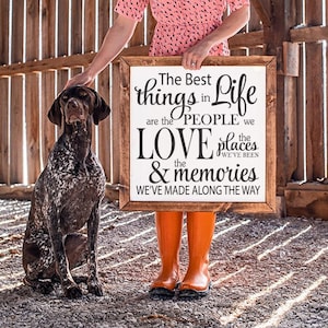 Op de afbeelding: Een houten bord met de tekst "The Best things in Life are the PEOPLE we LOVE the places & memories WE'VE MADE ALONG THE WAY." Een bruin-witte hond zit naast het bord. Het bord wordt vastgehouden door een persoon met oranje laarzen.