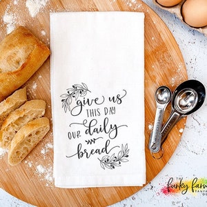 May include: A white kitchen towel with the text "give us this day our daily bread" sits on a wooden cutting board with a loaf of bread and measuring spoons. A carton of eggs is in the background.