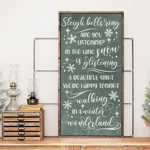 May include: A framed chalkboard sign with a green background and white lettering that reads "Sleigh bells ring...walking in a winter wonderland." The sign is decorated with snowflakes and stars. Two small Christmas trees and a wooden drawer unit are on a wooden surface.
