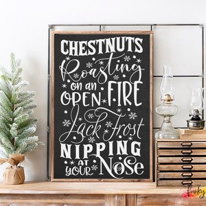 May include: A framed chalkboard sign with the words "Chestnuts Roasting on an Open Fire, Jack Frost Nipping at Your Nose" in white script. Two small Christmas trees flank the sign. Decorative lamps and a wooden storage unit are also visible.