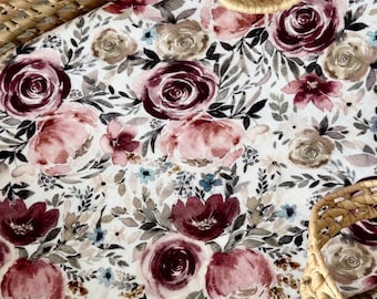 May include: A wicker bassinet with a floral patterned insert. The insert has a white background with burgundy, pink, and tan roses, and green and blue foliage. The bassinet has two woven handles.