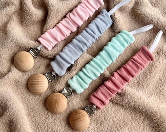 Può includere: Quattro clip per ciuccio per bambini in rosa, grigio, verde menta e rosso-rosa. Ogni clip ha un disco di legno e una fascia elastica rivestita in tessuto. Le clip sono disposte su una morbida coperta beige.