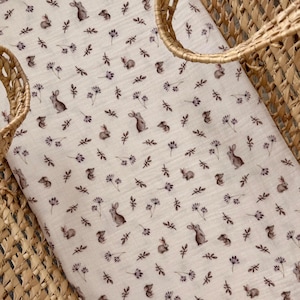 May include: A cream-colored baby bassinet sheet with a repeating pattern of small brown bunnies and purple floral accents. The sheet is nestled inside a woven wicker bassinet.