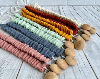 May include: A collection of baby pacifier clips in various colors, including pink, blue, green, yellow, orange, and cream. Each clip features a fabric strap with a gathered texture and a wooden clip. White ribbon loops are attached to the fabric.