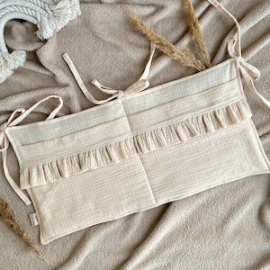 May include: A beige crib bumper with a ruffled edge and ties. The bumper is made of a soft, breathable fabric and is designed to protect babies from bumps and bruises.