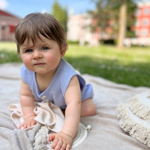 May include: A baby wearing a light blue sleeveless onesie sits on a cream-colored blanket. The baby is looking at the camera with a serious expression. The blanket has a fringe trim and is on a grassy surface.