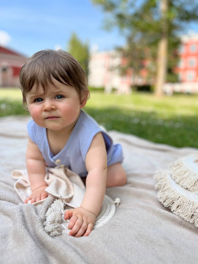 May include: A baby wearing a light blue sleeveless onesie sits on a cream-colored blanket. The baby is looking at the camera with a serious expression. The blanket has a fringe trim and is on a grassy surface.