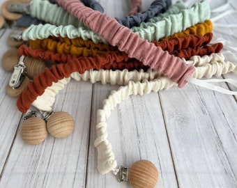 May include: A collection of baby pacifier clips in various colors, including cream, rust, mustard, and dusty rose. Each clip features a wooden bead and a metal clasp. The fabric is gathered and textured.