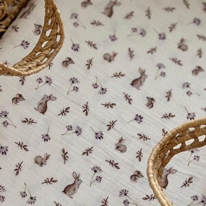 May include: A white baby blanket with a repeating pattern of brown bunnies, small flowers, and sprigs. The blanket is partially framed by a woven basket. The overall design is soft and delicate, suitable for a nursery.