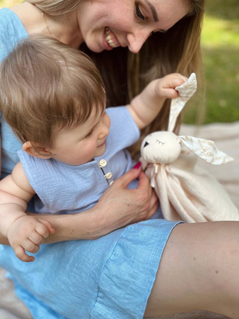 May include: A woman in a light blue dress holds a baby in her lap. The baby is holding a soft, white bunny toy with embroidered eyes and a gold floral pattern. The woman is smiling at the baby.