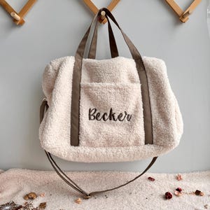 Pode incluir: Uma bolsa tote creme e fofa com alças marrons e uma alça de ombro combinando. A palavra "Becker" é bordada em uma fonte cursiva no bolso frontal. A bolsa tem uma aparência macia e texturizada.