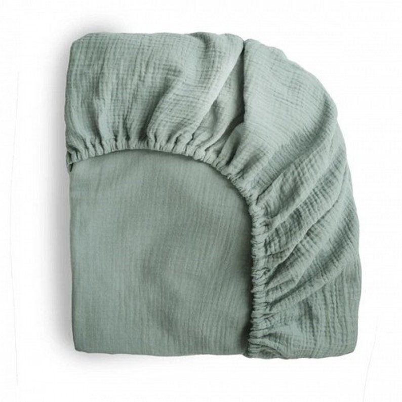 May include: A sage green fitted sheet with a textured, crinkled appearance. The sheet is made of a soft fabric and has elastic edges for a snug fit. The sheet is designed to fit a mattress and is shown on a white background.