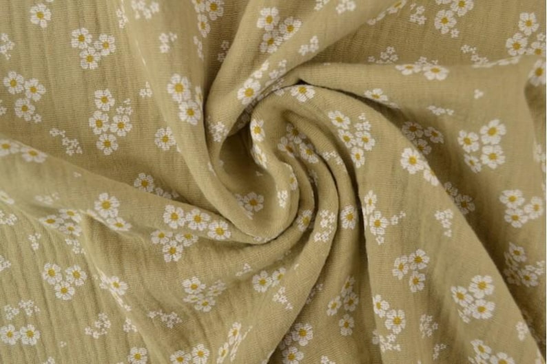 May include: Close-up of a fabric with a repeating floral pattern. The fabric is a light olive green color, with small white flowers and yellow centers. The fabric is soft and flowing, with visible texture.
