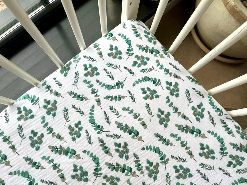 May include: White fitted crib sheet with a repeating pattern of green and gray eucalyptus leaves. The sheet is made of a soft, textured fabric and is placed on a white crib.