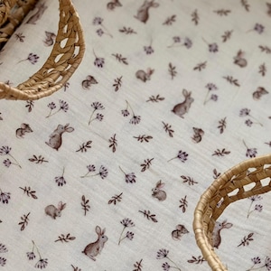 Op de afbeelding: Een witte babydeken met een herhalend patroon van bruine konijntjes, kleine bloemen en bladeren. Een gevlochten mand is op de voorgrond te zien. De deken is gemaakt van een zachte, lichte stof.