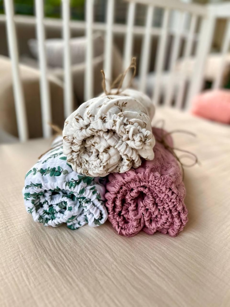 May include: Three rolled up baby swaddles in different patterns. One is white with brown floral print, one is green with white leaves, and one is pink with white floral print.