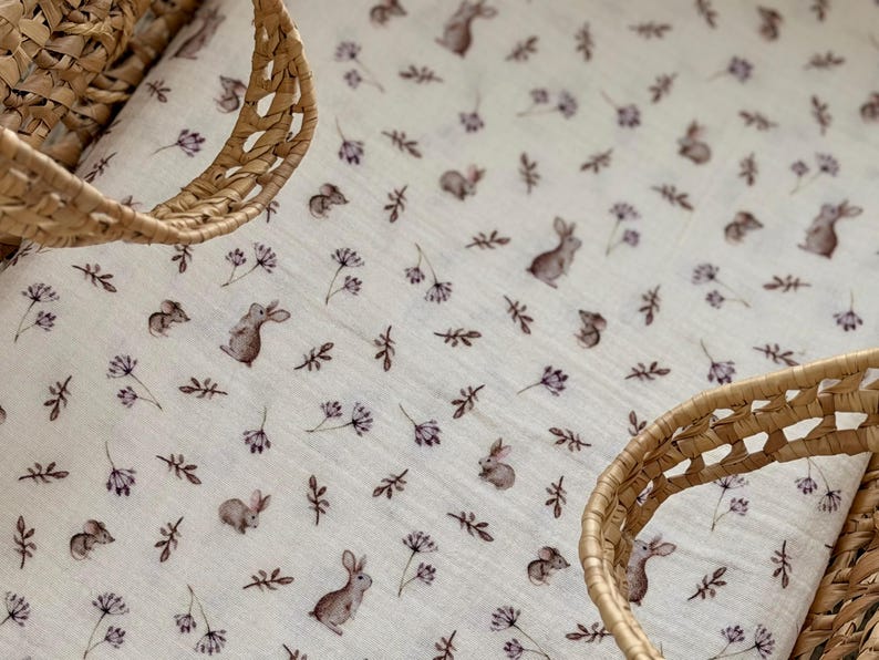 May include: A white baby blanket with a repeating pattern of brown bunnies, purple flowers, and green leaves. The blanket is partially framed by a woven wicker basket. The overall design is soft and delicate, suitable for a nursery.