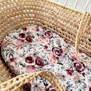 May include: Woven wicker bassinet with a floral patterned mattress pad. The pad features a white background with burgundy, pink, and gray roses, along with blue and green foliage. The bassinet has a sturdy, open weave design.