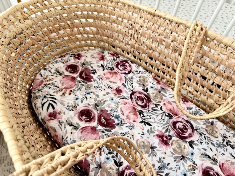 May include: Woven wicker bassinet with a floral patterned mattress pad. The pad features a white background with burgundy, pink, and gray roses, along with blue and green foliage. The bassinet has a sturdy, open weave design.