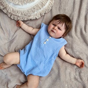 May include: A baby wearing a light blue sleeveless onesie with button closures. The baby is lying on a light brown fuzzy blanket.