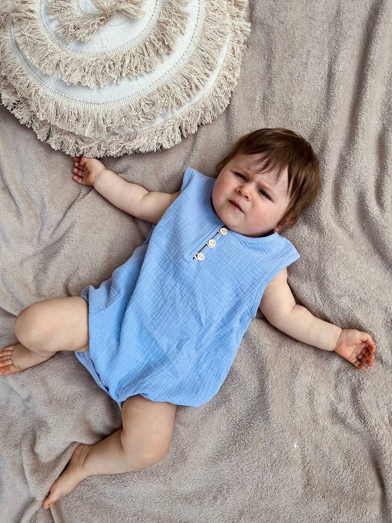 May include: A baby wearing a light blue sleeveless onesie with button closures. The baby is lying on a light brown fuzzy blanket.