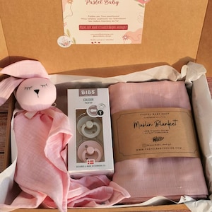 K&ouml;nnte beinhalten: Eine rosa Geschenkbox gef&uuml;llt mit Babyartikeln. Die Box enth&auml;lt eine rosa Musselin-Decke, eine rosa Hasenrassel und eine Packung BIBS-Schnuller. Die Box ist mit "Pastel Baby Shop" und "Muslin Blanket" beschriftet.