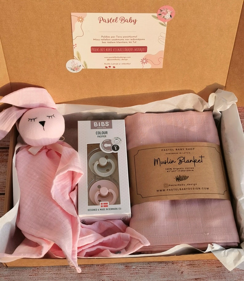 K&ouml;nnte beinhalten: Eine rosa Geschenkbox gef&uuml;llt mit Babyartikeln. Die Box enth&auml;lt eine rosa Musselin-Decke, eine rosa Hasenrassel und eine Packung BIBS-Schnuller. Die Box ist mit "Pastel Baby Shop" und "Muslin Blanket" beschriftet.