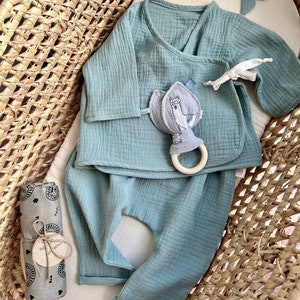 May include: A baby outfit set in a light blue color, including a long-sleeved shirt with a wrap-style neckline and matching pants. The set is laid out in a woven basket with a small stuffed animal and a wooden ring.