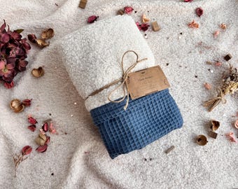 May include: A folded blanket with a white, fluffy top and a blue, textured bottom. The blanket is tied with twine and has a brown tag attached. The blanket is on a soft, white surface with decorative elements.