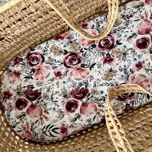 Puede incluir: Una cuna de mimbre con un inserto con estampado floral. El inserto presenta un fondo blanco con rosas burdeos, rosas y beige, junto con follaje verde y azul. La cuna tiene dos asas tejidas.