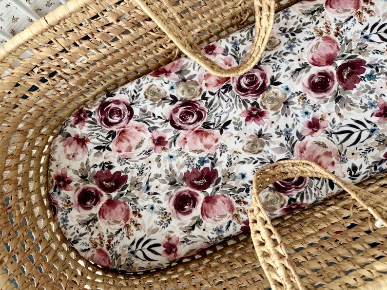 Puede incluir: Una cuna de mimbre con un inserto con estampado floral. El inserto presenta un fondo blanco con rosas burdeos, rosas y beige, junto con follaje verde y azul. La cuna tiene dos asas tejidas.