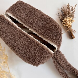 May include: A brown, rectangular, fluffy zippered pouch with a white zipper and pull. The pouch is made of a soft, textured material. A small bundle of dried flowers is next to the pouch.