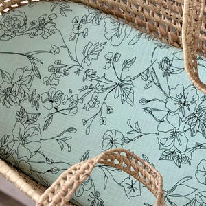 May include: A light blue baby bassinet sheet with a floral pattern. The sheet features black line drawings of flowers and leaves. The bassinet is made of woven wicker.