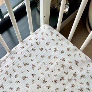 May include: A white fitted crib sheet with a repeating pattern of small brown leaves. The sheet is made of a soft, textured fabric and is placed inside a white crib with rounded spindles.