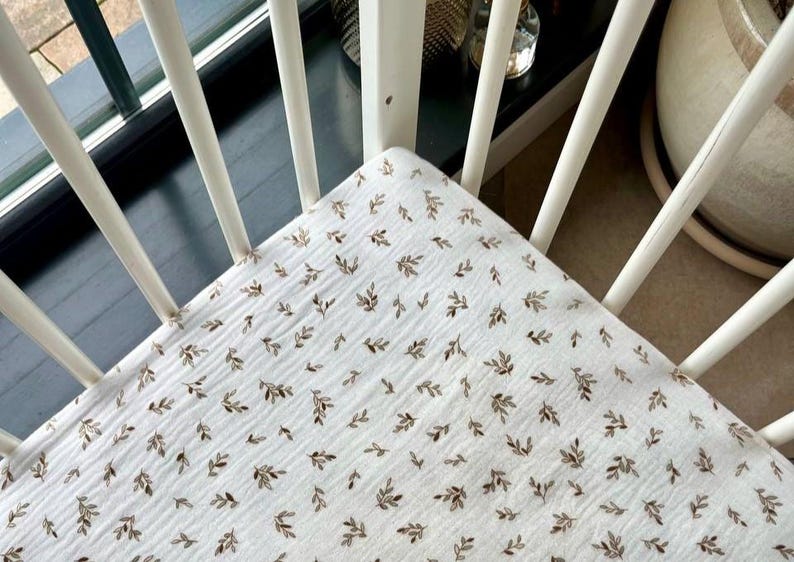 May include: A white fitted crib sheet with a repeating pattern of small brown leaves. The sheet is made of a soft, textured fabric and is placed inside a white crib with rounded spindles.