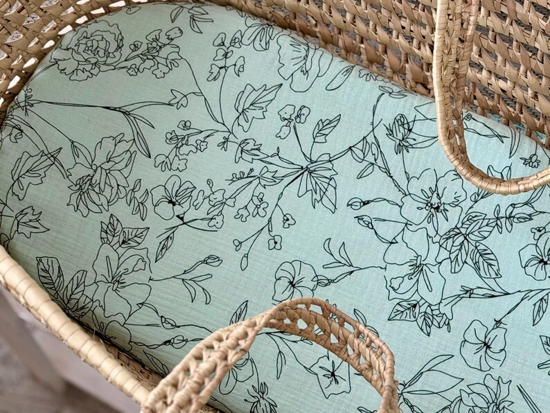 May include: A light blue baby bassinet sheet with a floral pattern. The sheet features black line drawings of flowers and leaves. The bassinet is made of woven wicker.